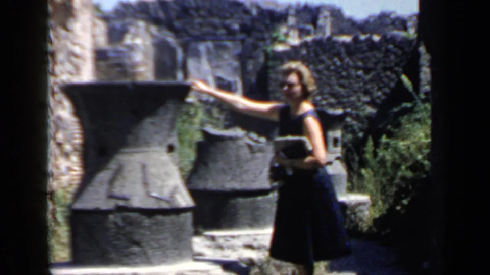 In Rome travel footage, a woman gestures toward a large stone vessel while holding a notebook at an ancient ruin site.