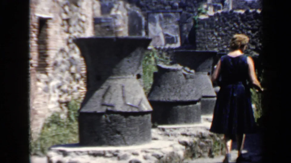 A woman in a dark dress walks away through a Roman ruin area with large stone vessels and weathered masonry.