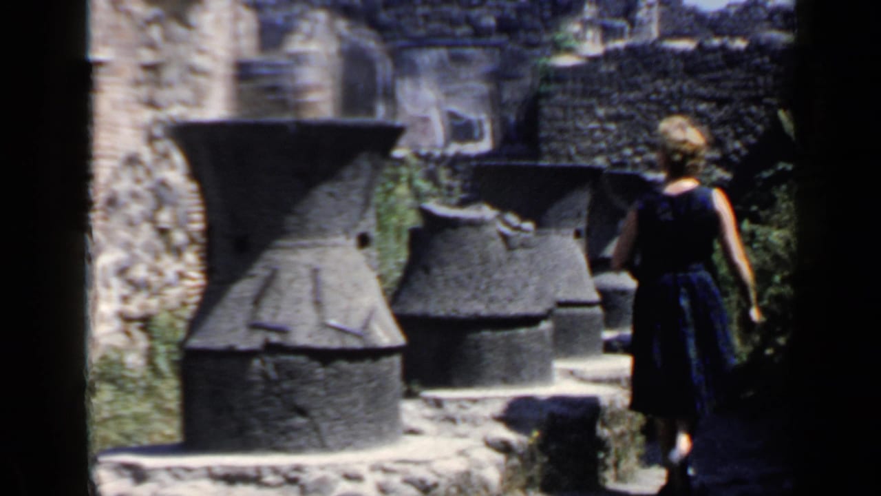 Woman in a dark dress walks past stone ruins seen from a shaded archway.
