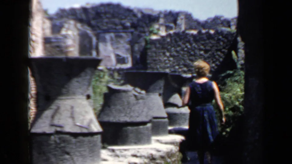 Woman in a dark dress pauses among weathered stone structures at an Italian ruins site in 1960s home-movie footage.