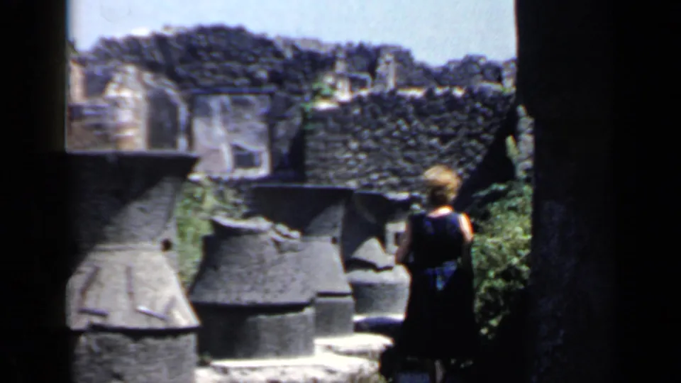 A woman walks away from the camera through sunlit ruins, with rough masonry and rounded stone forms filling the frame.