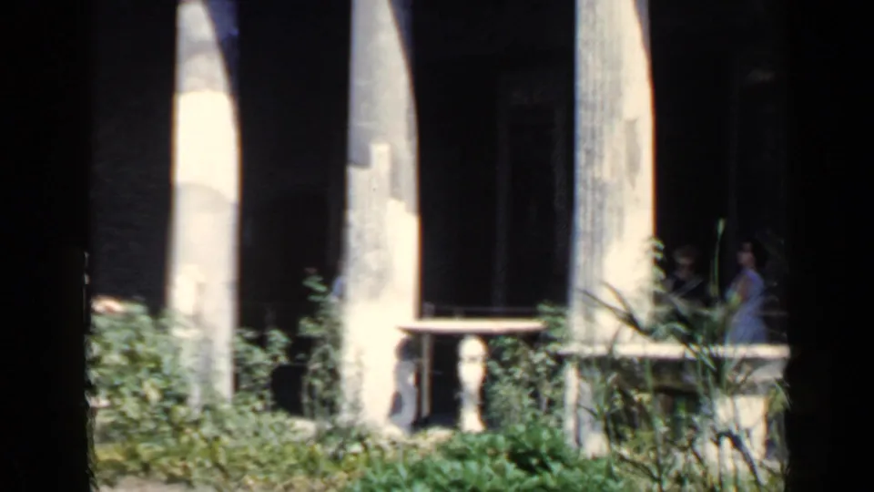 1960 Rome ruins frame showing pale columns, shadowed openings, greenery, railing, and a distant visitor near the right edge.