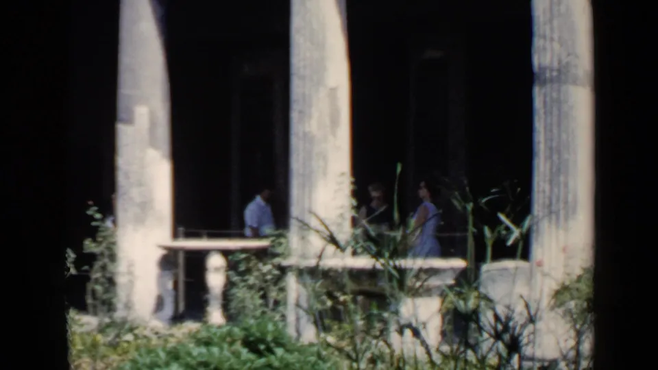 Tourists in summer clothing are visible among worn columns and greenery at a historic site in Rome, filmed with soft 8mm color.