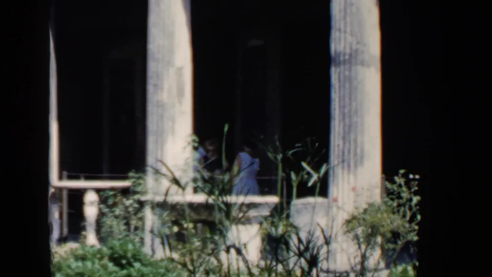 A woman in a light summer dress appears near overgrown plants and tall stone columns within shaded Roman ruin footage from 1960.