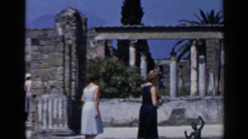 Two women in summer dresses walk through a sunlit historic stone area in Rome, with arches, walls, plants, and blue sky visible.
