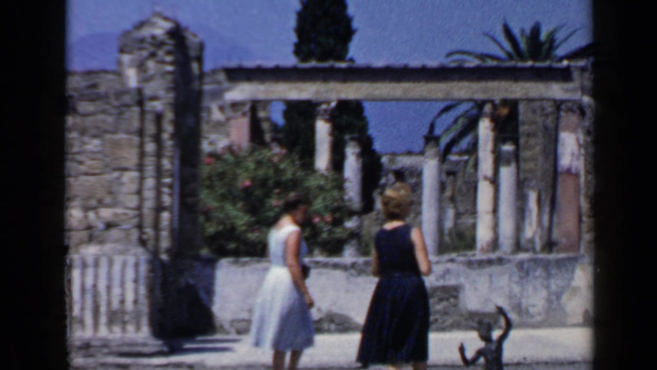 Two women walk past stone columns and a low wall at ancient ruins.