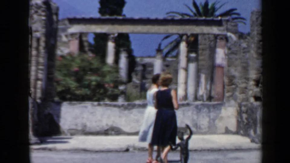 Two women in mid-century dresses move through a sunlit Italian ruin site, framed by columns, stonework, palms, and shrubs.