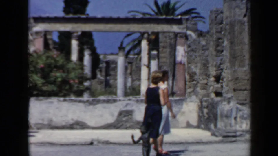 Two female tourists walk together through ancient ruins in Italy, with a low wall, columns, plants, and sunlit stonework visible.