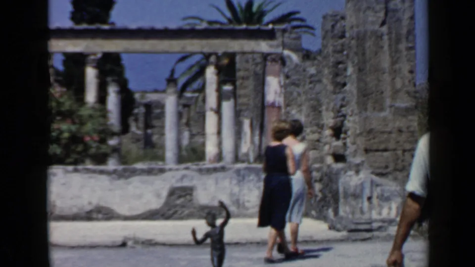 Two women walk through the ruin site while part of another adult enters at the right edge, with columns and stone walls in view.