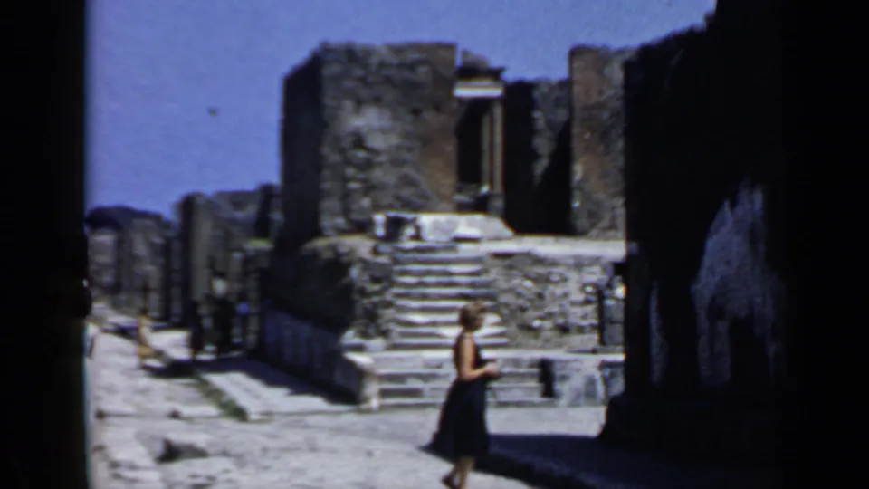 A tourist in a dark dress walks beside weathered stone steps and walls in the Pompeii ruins.