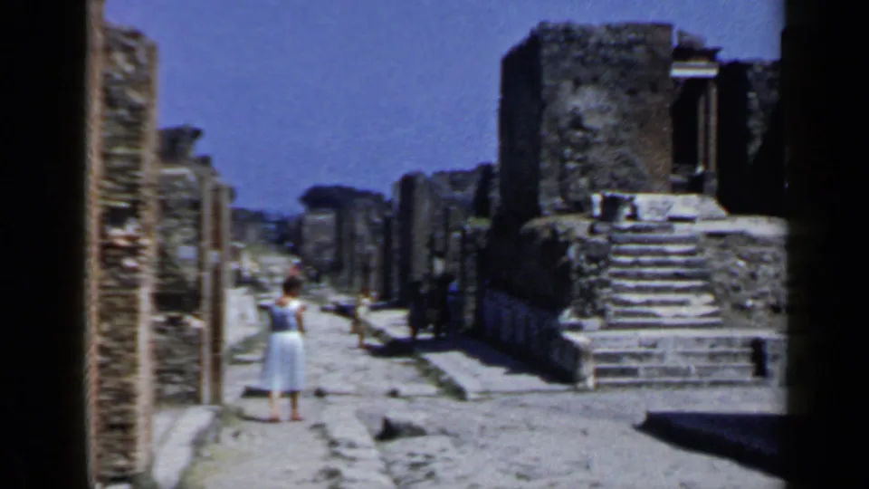 Tourists in light summer clothing appear on a stone street framed by ruins in 1960 Pompeii footage.
