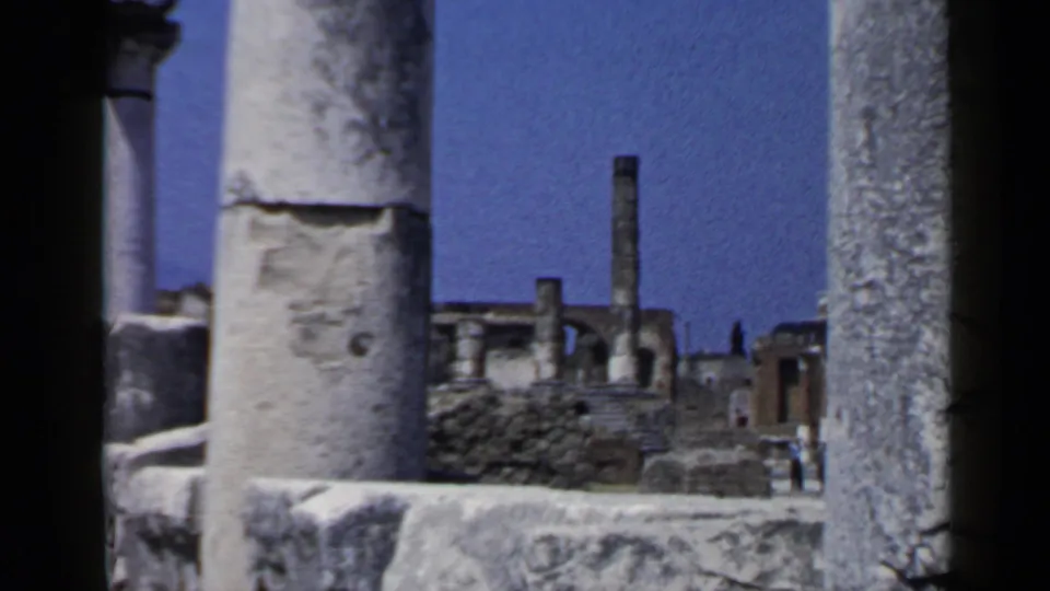 A 1960 8mm view of columns, low walls, and distant ruins at Pompeii under clear sky.