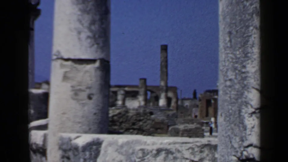 1960 8mm view of Pompeii ruins seen through weathered columns, with distant visitors among stone structures.