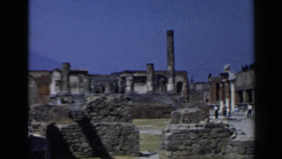 1960 home-movie frame of Pompeii with masonry walls, upright columns, and tourists walking through the ruins.