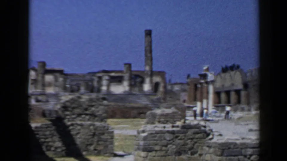 Wider 1960 8mm view of Pompeii ruins, with low masonry, standing columns, and visitors crossing the archaeological grounds.