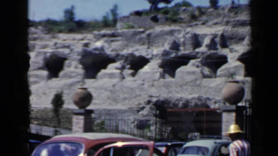 At the Rome ruins, a red mid-century car, other parked vehicles, fencing, pottery-like vessels, and a sunhat-wearing visitor appear in the foreground.