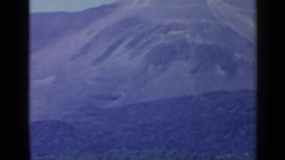 Faded 8mm frame of a hazy mountain ridge and lower hillside, tinted blue by the aged film color.