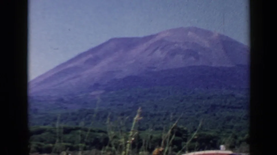 1960 8mm home-movie frame of a large mountain above forest, with roadside grasses in the foreground.