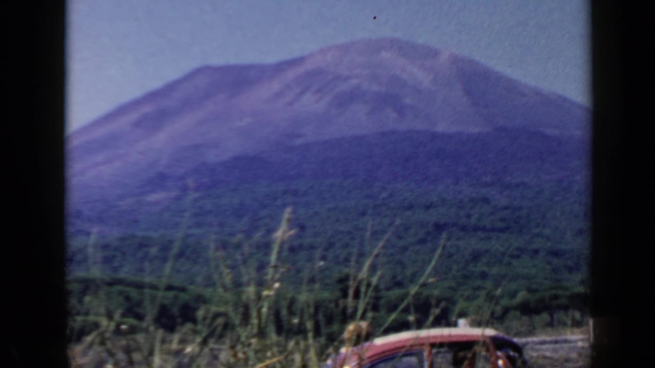 1960 8mm Rome-area travel frame showing a broad mountain beyond forest, with roadside grasses and a partial red car visible below.