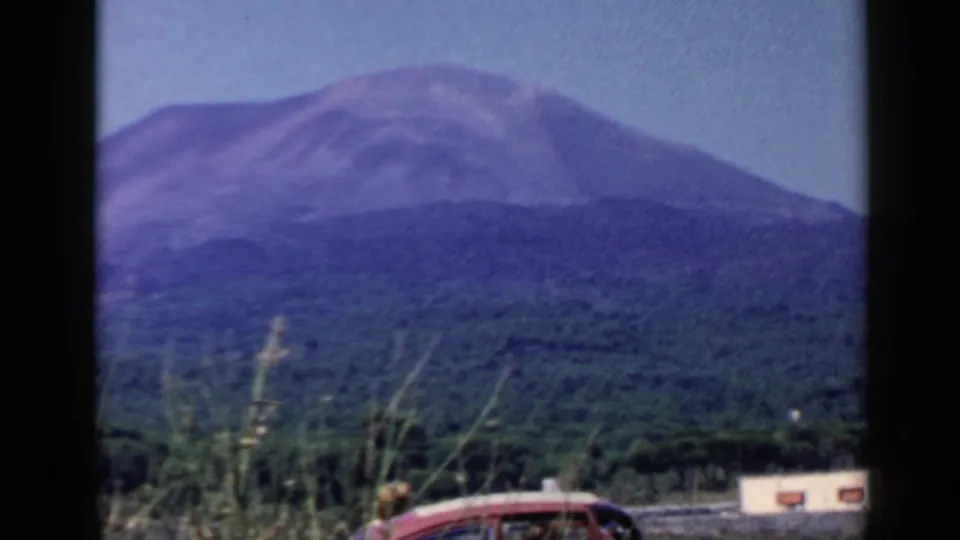 1960 8mm Rome-area landscape view with a mountain over forest, a partial red car, roadside grass, and a small pale building at right.