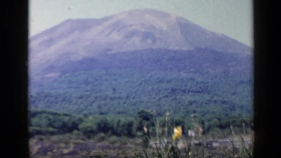 1960 8mm Rome-area frame with a softly exposed mountain, green forest, pale sky, and foreground grasses along the roadside.
