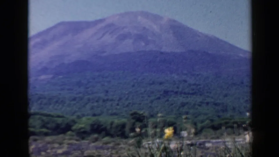1960 8mm Rome-area travel frame showing the mountain and forest more tightly, with dark gate edges and roadside vegetation below.