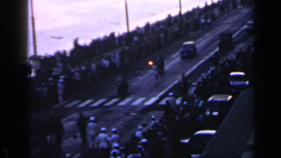 Rome 1960 Olympic parade street scene with a crowd near a crosswalk, parked cars, and water beyond the avenue.
