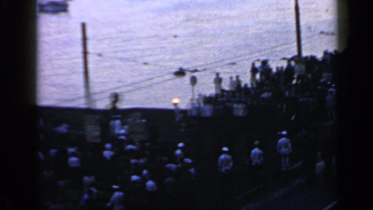 Crowds line a dark riverside walkway as a small torch flame glows near the water.