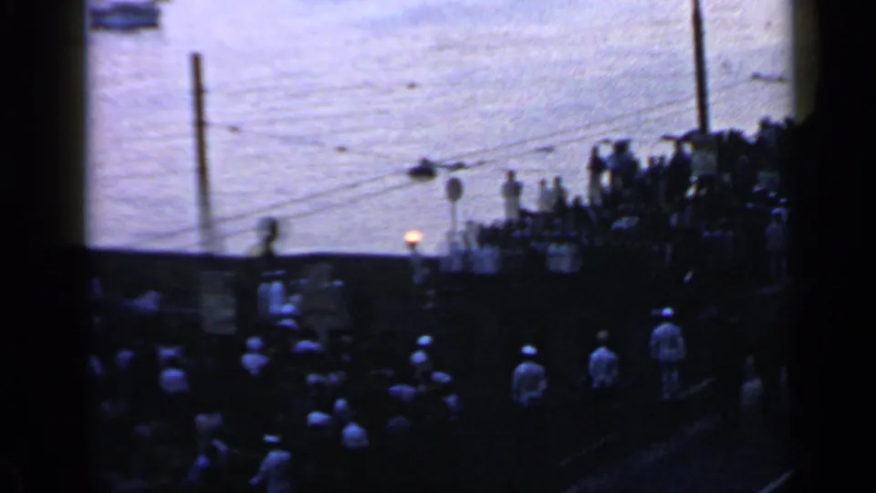 1960 Rome home-movie frame showing spectators gathered along a riverside promenade with a torch flame visible near the water at dusk.