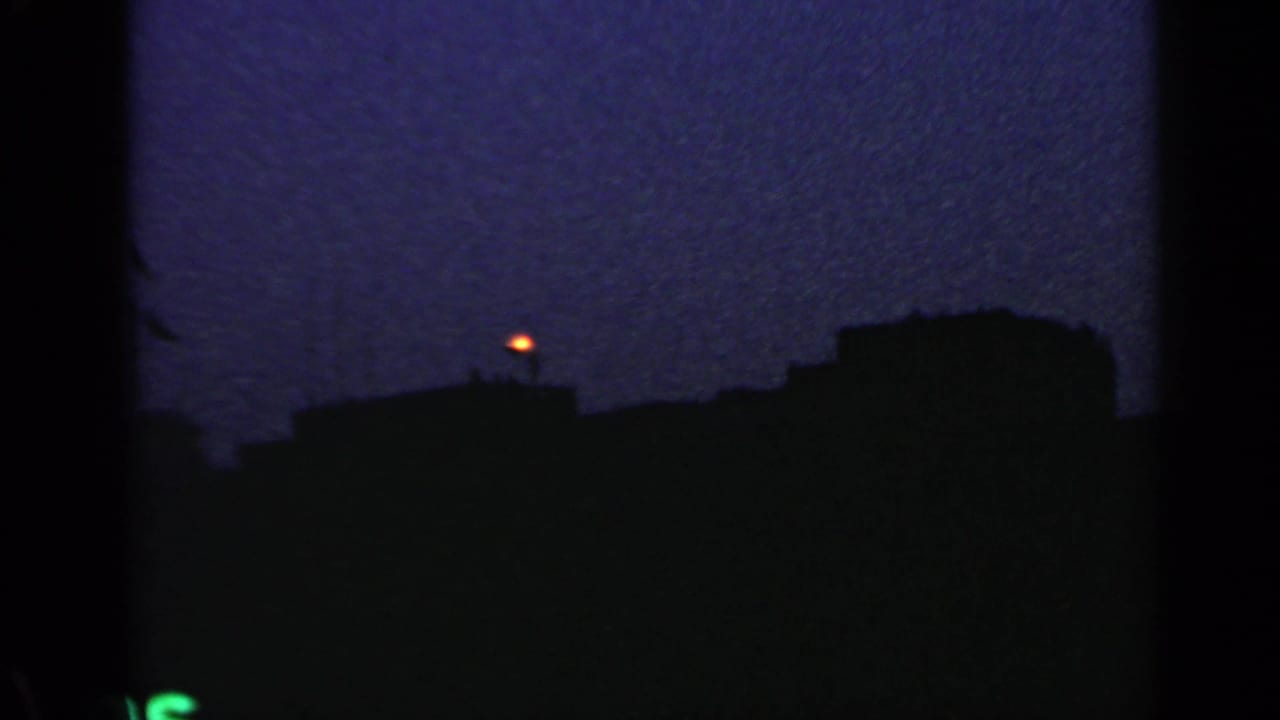 Dark rooftops silhouetted beneath a grainy blue evening sky with a small orange light above the skyline.