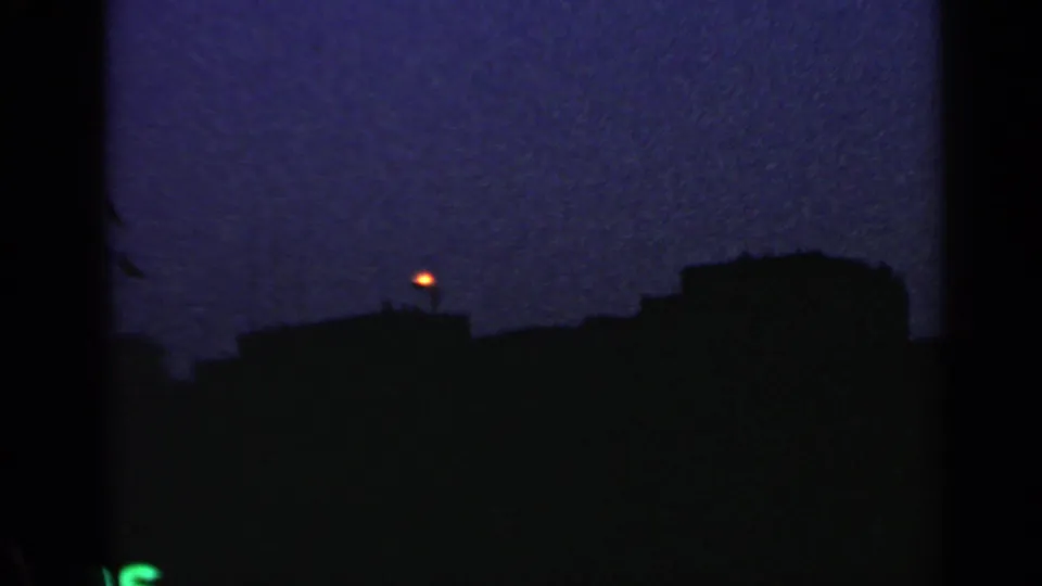 1960 Rome home-movie frame showing silhouetted rooftops and a small orange light in a dark, grainy evening sky.