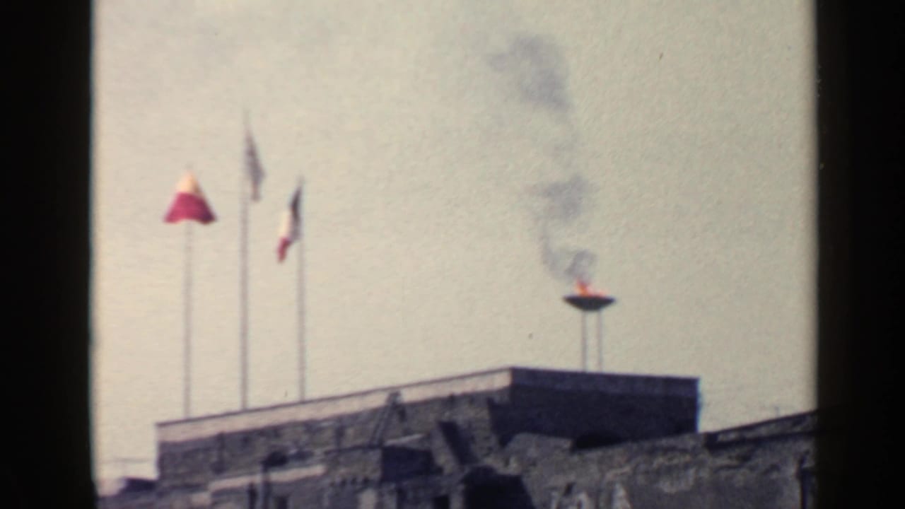 Three flags and a smoking flame above a stone rooftop against a pale sky.