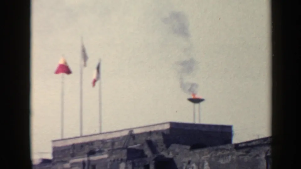 1960 Rome 8mm frame showing three flags beside a raised flame bowl sending dark smoke above a stone rooftop.