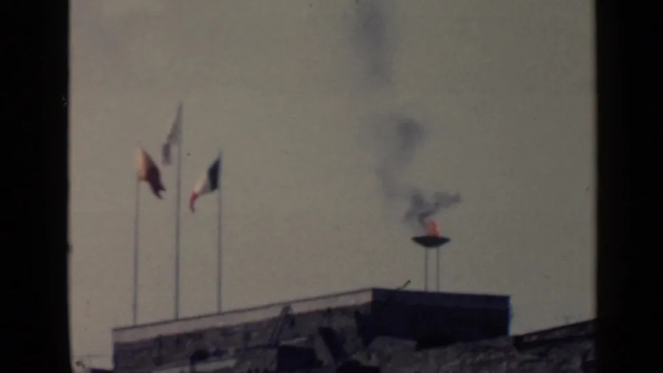 1960 Rome home-movie frame with three flags, a small flame, and black smoke rising from a bowl near a stone parapet.