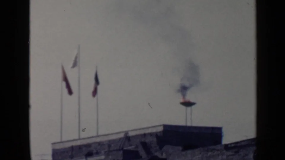 1960 Rome 8mm frame with a raised fire bowl, dark smoke, and three flags above a stone wall under a pale sky.
