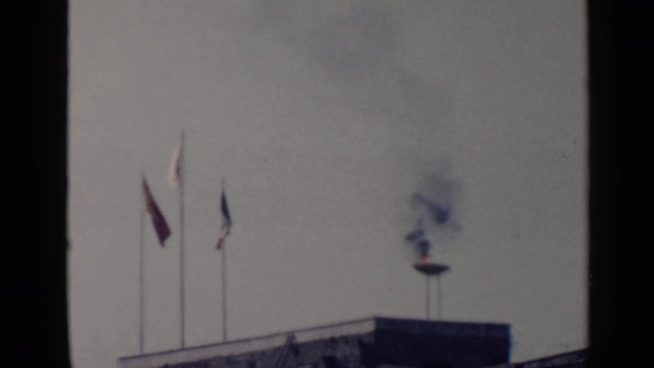 Three flags on poles beside a rooftop flame emitting smoke against a pale sky.