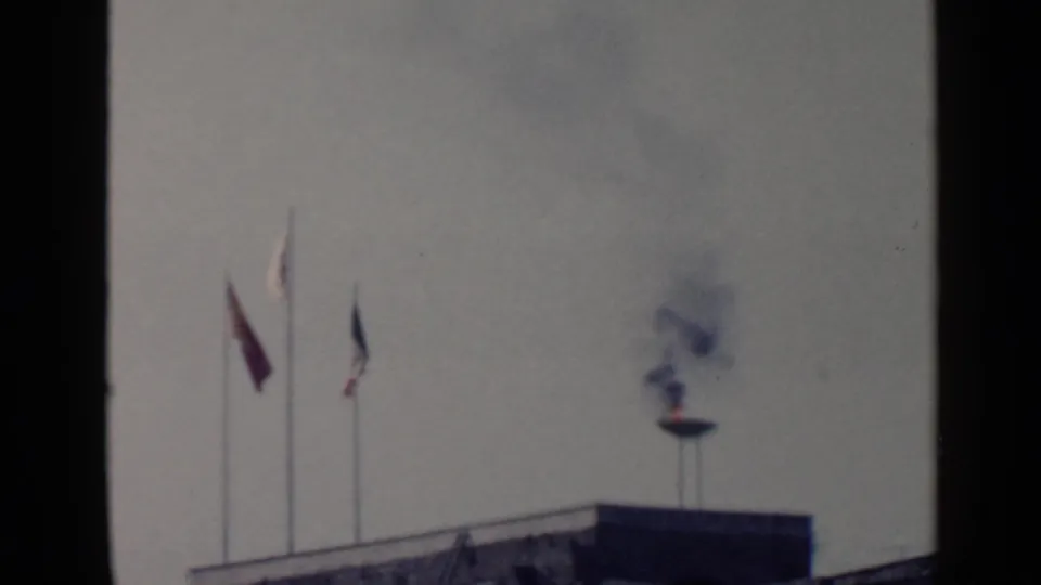 1960 Rome home-movie frame showing three flagpoles and a smoking flame bowl above a building edge.