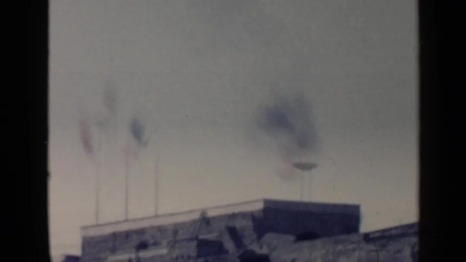 Soft 8mm frame from Rome showing flags and a smoky flame on or above a rooftop structure.