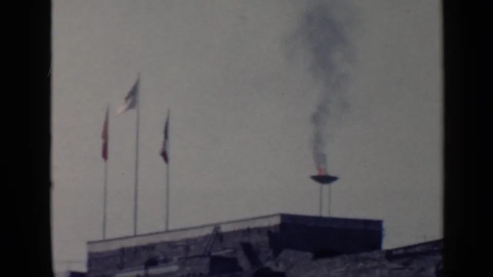 1960 Rome 8mm frame with a rooftop or terrace flame sending smoke upward beside raised flags.