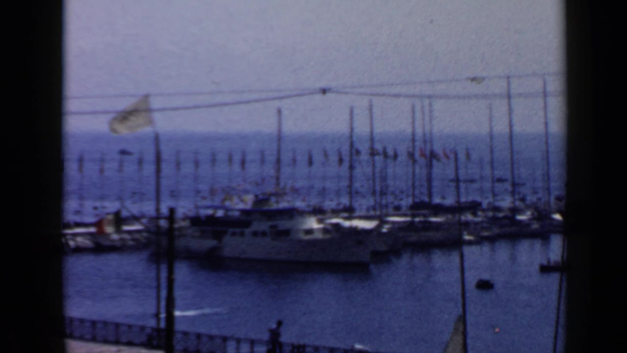 1960 Rome home-movie frame showing a marina through dark window framing, with yachts, flags, and calm blue harbor water.
