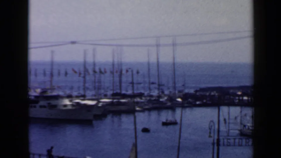 1960 Rome marina frame showing sailboat masts, a line of flags, calm harbor water, and a partial restaurant sign at the edge.