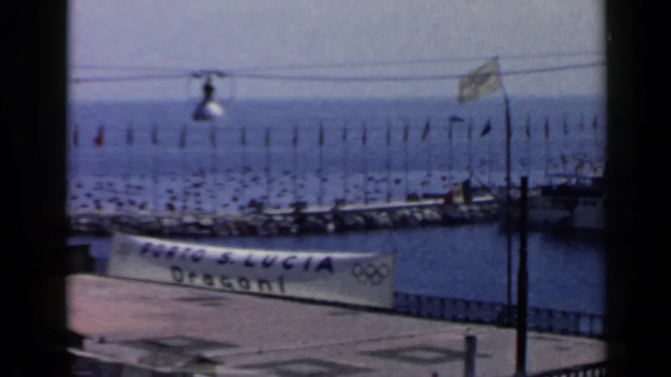 1960 Rome-area marina frame showing a Porto S. Lucia Dragoni banner with Olympic rings, flags, lamp post and boats.