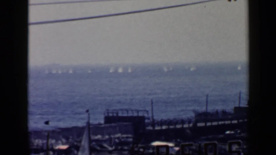 Blue-tinted 1960 Rome-area coastal view with distant sailboats on open water and marina structures in the foreground.