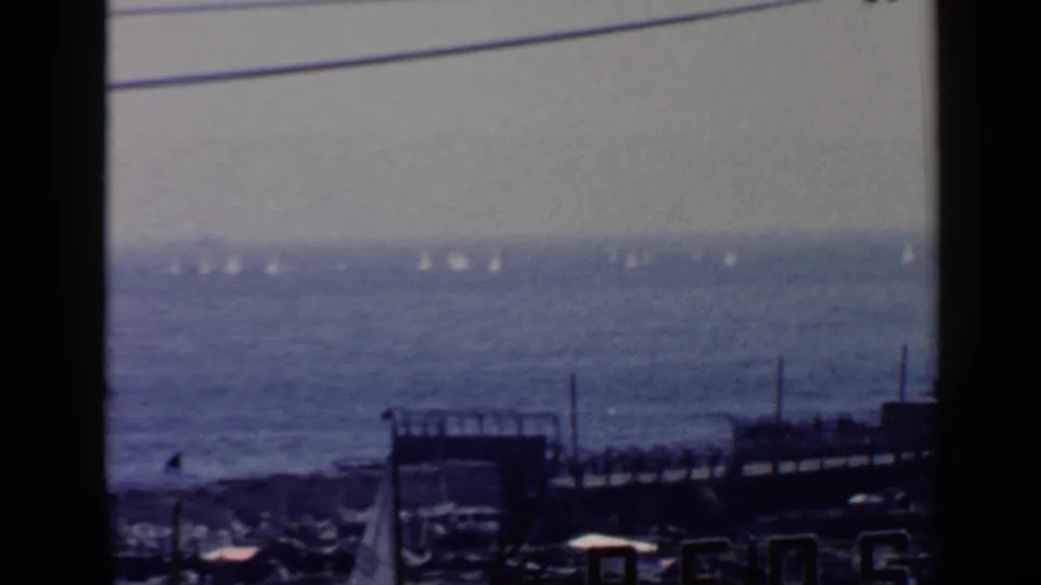 1960 Rome coastal home-movie frame showing small sailboats offshore, a pier-like structure, utility wires, and hazy sky.