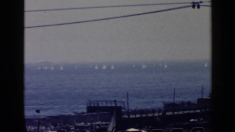 1960 Rome coastal view with sailboats on the Mediterranean, a dark pier or dock, overhead wires, and a hazy horizon.