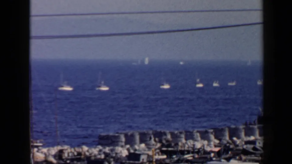 1960 Rome coastal home-movie frame with multiple sailboats offshore, rocky waterfront details, overhead wires, and a blue hazy sea.