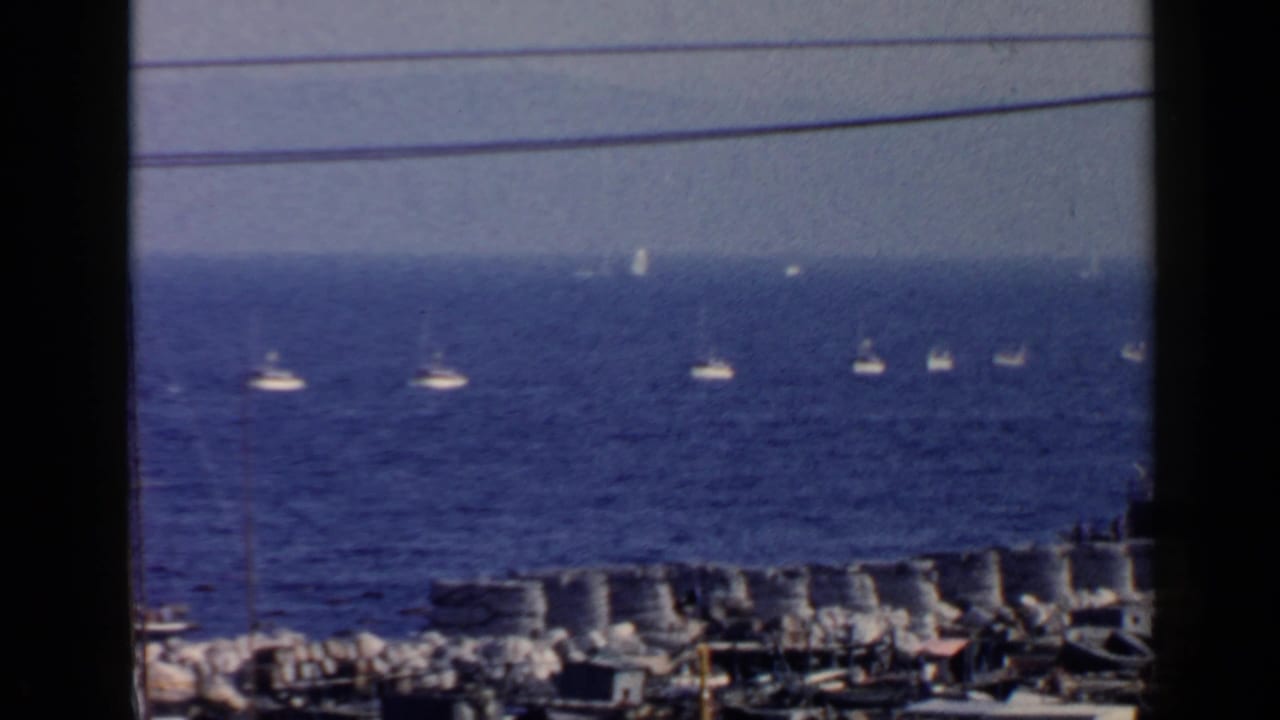 Blue bay with distant small boats, shoreline rocks, utility lines, and dark film-frame edges.