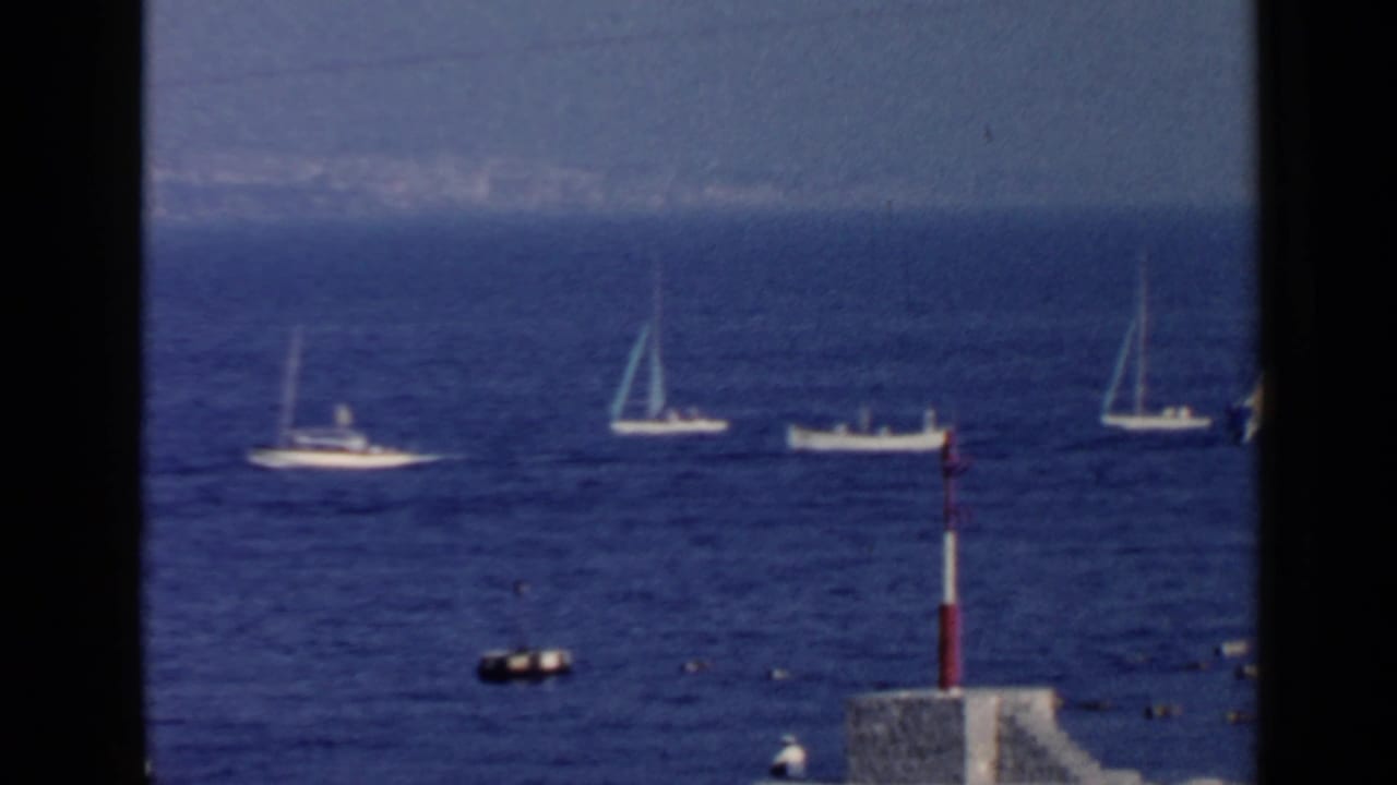 1960 8mm Rome coastal view with sailboats and a small motorboat on the Mediterranean, framed by dark vertical edges.