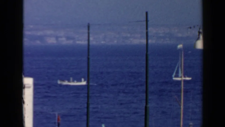 Rome 1960 8mm frame with distant boats on the Mediterranean, dark vertical foreground poles, and a partial sailboat mast at right.