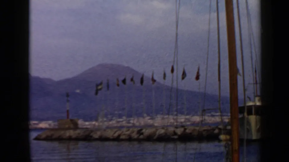 1960 8mm harbor view in Italy with flagpoles on a breakwater, sailboat rigging, calm water, and mountains in the distance.